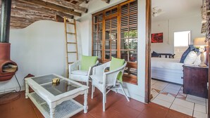 A room with a white wicker sofa set, a glass-top table, a wooden ladder, and a view through large wooden shutters to a bed and a lamp. - HoneyPot Guesthouse (Umhlanga)