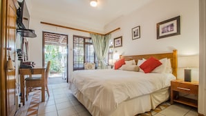 A bedroom with a large bed, a desk, and a chair. There is a window with curtains and a view of greenery outside. - HoneyPot Guesthouse (Umhlanga)
