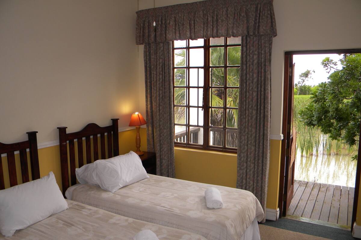 A hotel room with two beds, a window with curtains, a lamp, and a view of a water body outside.