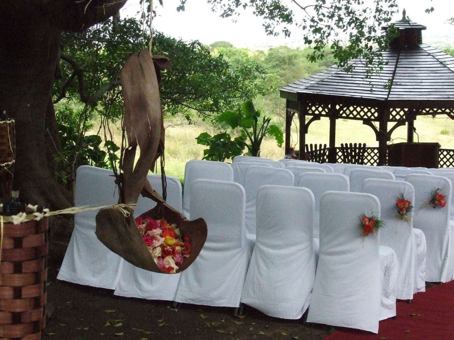 A gazebo with a wooden roof and lattice design, surrounded by white chairs with white tablecloths and floral centerpieces.