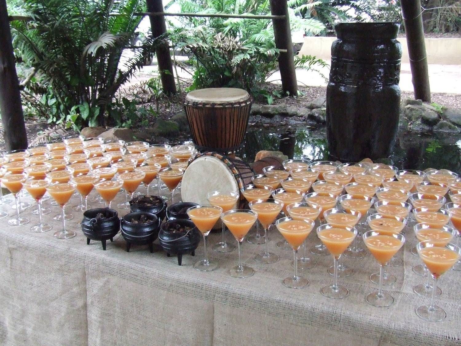 Une table dressée avec diverses boissons et de petits bols, entourée de plantes tropicales et d’une grande structure en pierre.