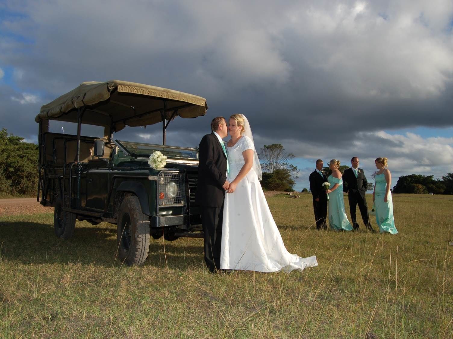 A wedding scene with a couple in front of a green vehicle, surrounded by guests in a grassy field.