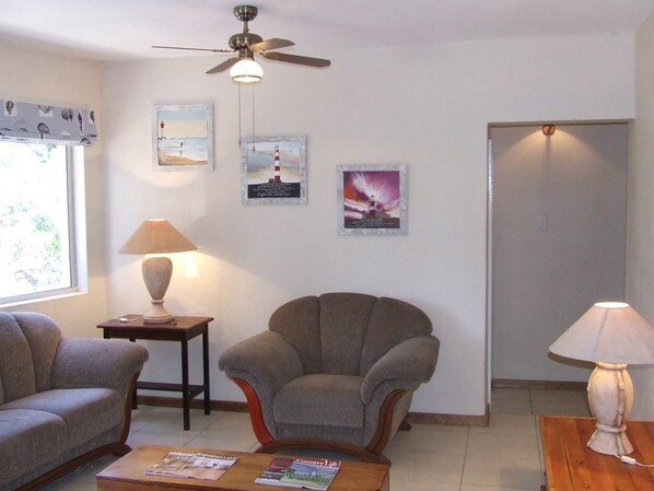 A living room with a sofa, armchair, coffee table, lamp, and ceiling fan. There are three framed pictures on the wall.