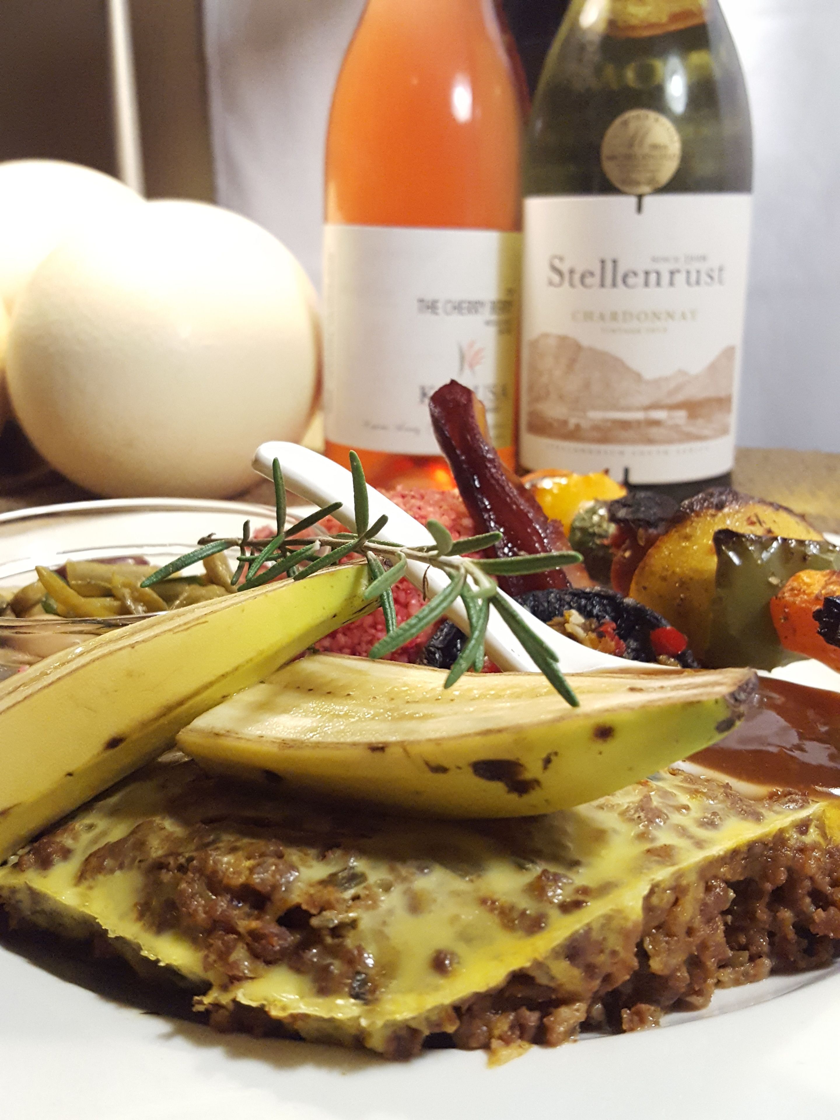 A plate of food with a slice of meatloaf topped with cheese and vegetables, a bottle of wine, and a roasted egg in the background.