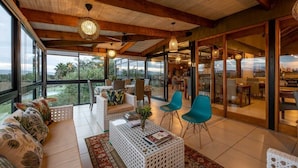 A spacious living area with a large sofa, a coffee table, and blue chairs. There are large windows offering a view of the outdoors. - De Zeekoe Guest Farm (Oudtshoorn)