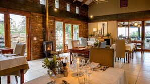 A dining area with tables set for a meal, a fireplace, and large windows. - De Zeekoe Guest Farm (Oudtshoorn)