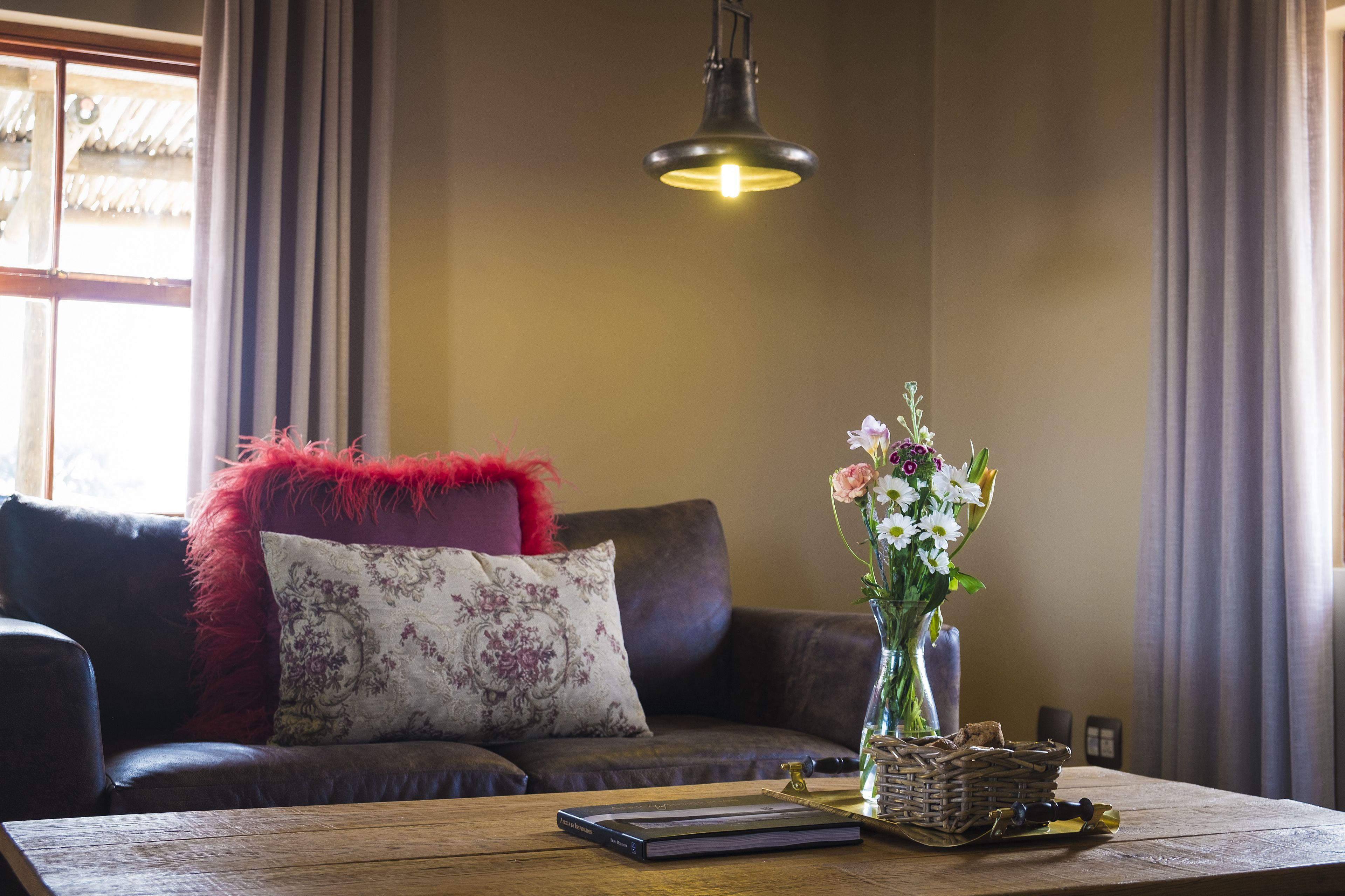 Uma sala de estar acolhedora com um sofá de couro escuro, um travesseiro vermelho, uma almofada floral, um vaso com flores e uma mesa de centro de madeira com um livro e uma cesta.