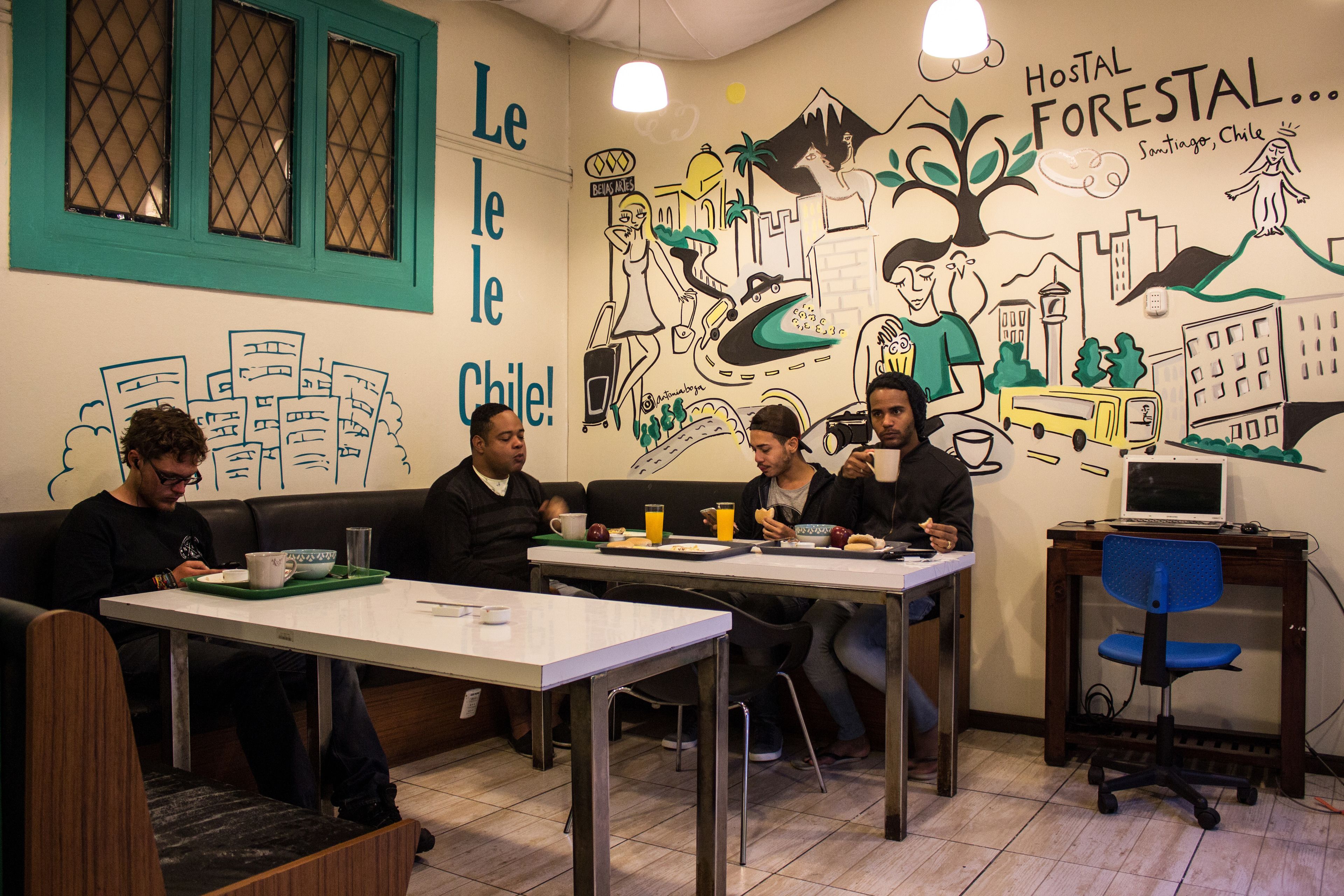 Un comedor con mesas y sillas, una laptop sobre un escritorio y un mural en la pared que representa diferentes escenas y texto.