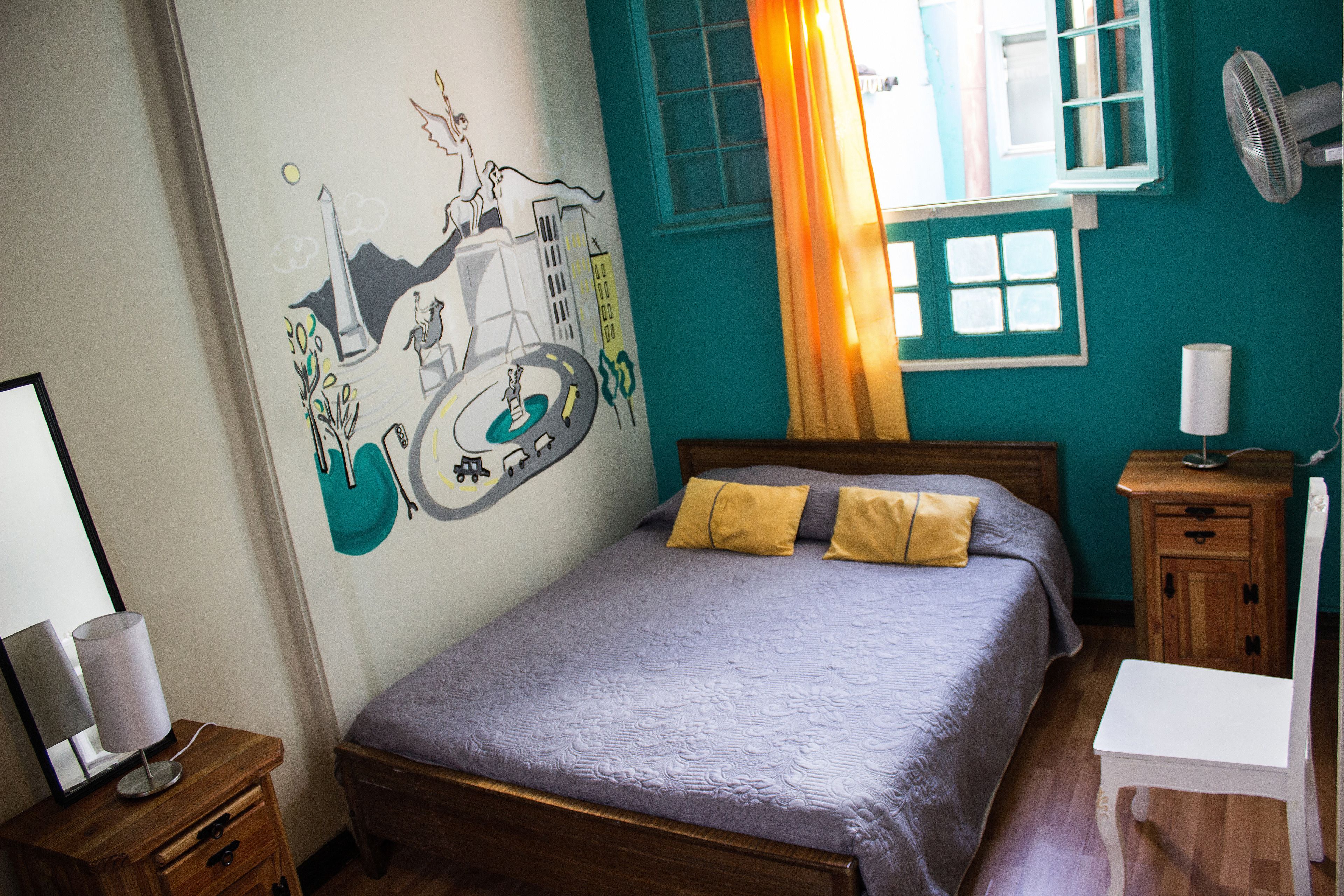 A bedroom with a bed, a nightstand, a lamp, a wall mural, and a window with curtains.