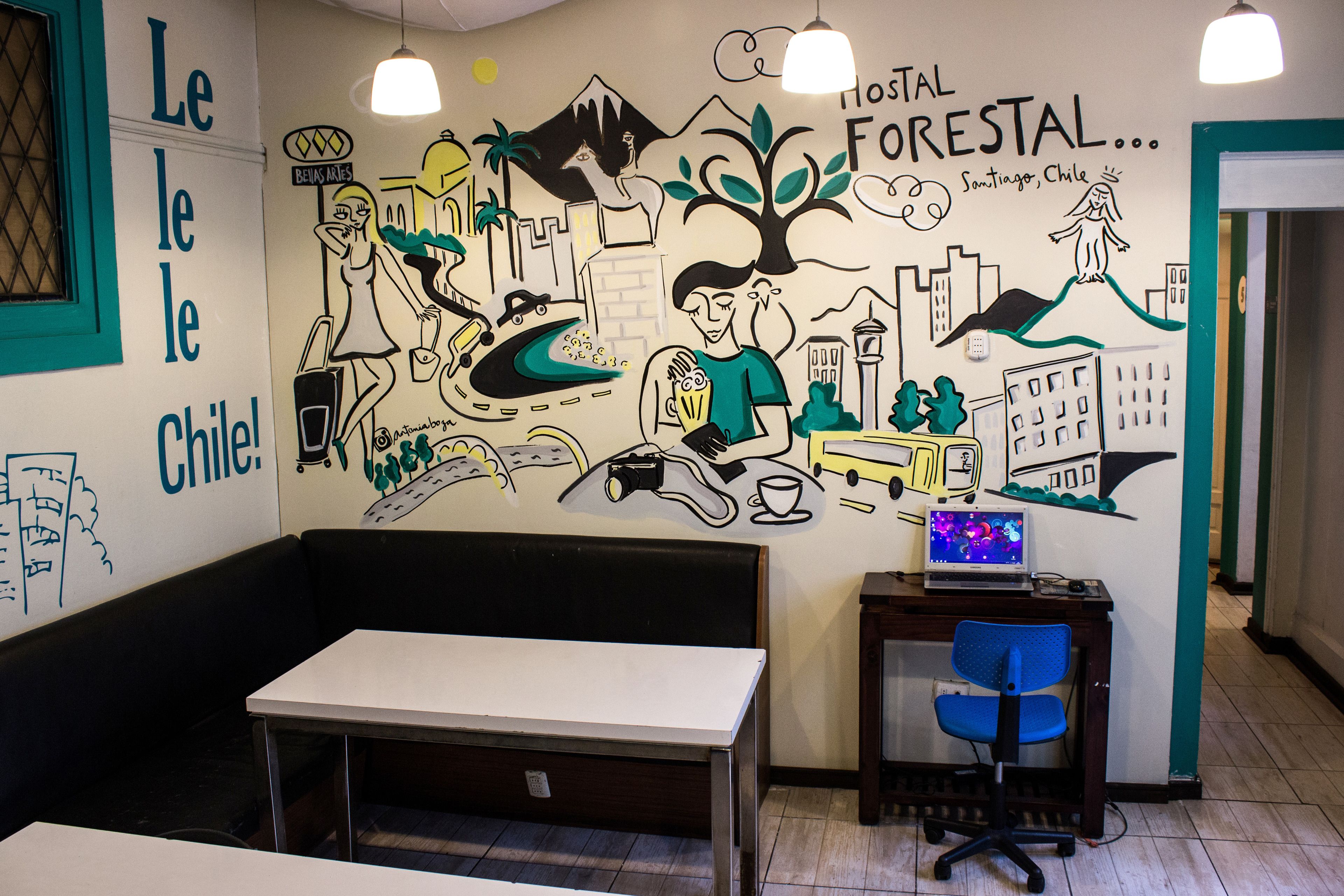 A room with a mural depicting Chilean scenes, a white table, a blue chair, and a laptop on a small table.