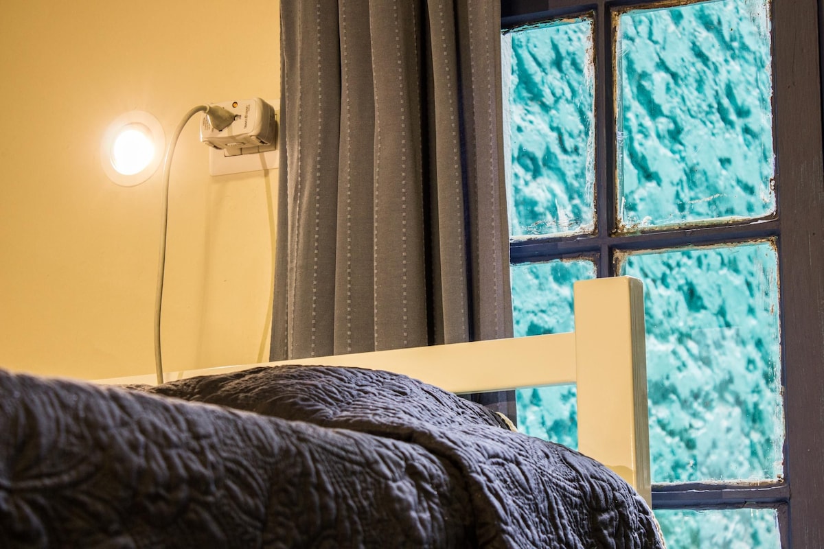 Una cama con un edredón oscuro y texturizado, una lámpara de noche, una ventana con vista a un cielo azul y un enchufe en la pared.