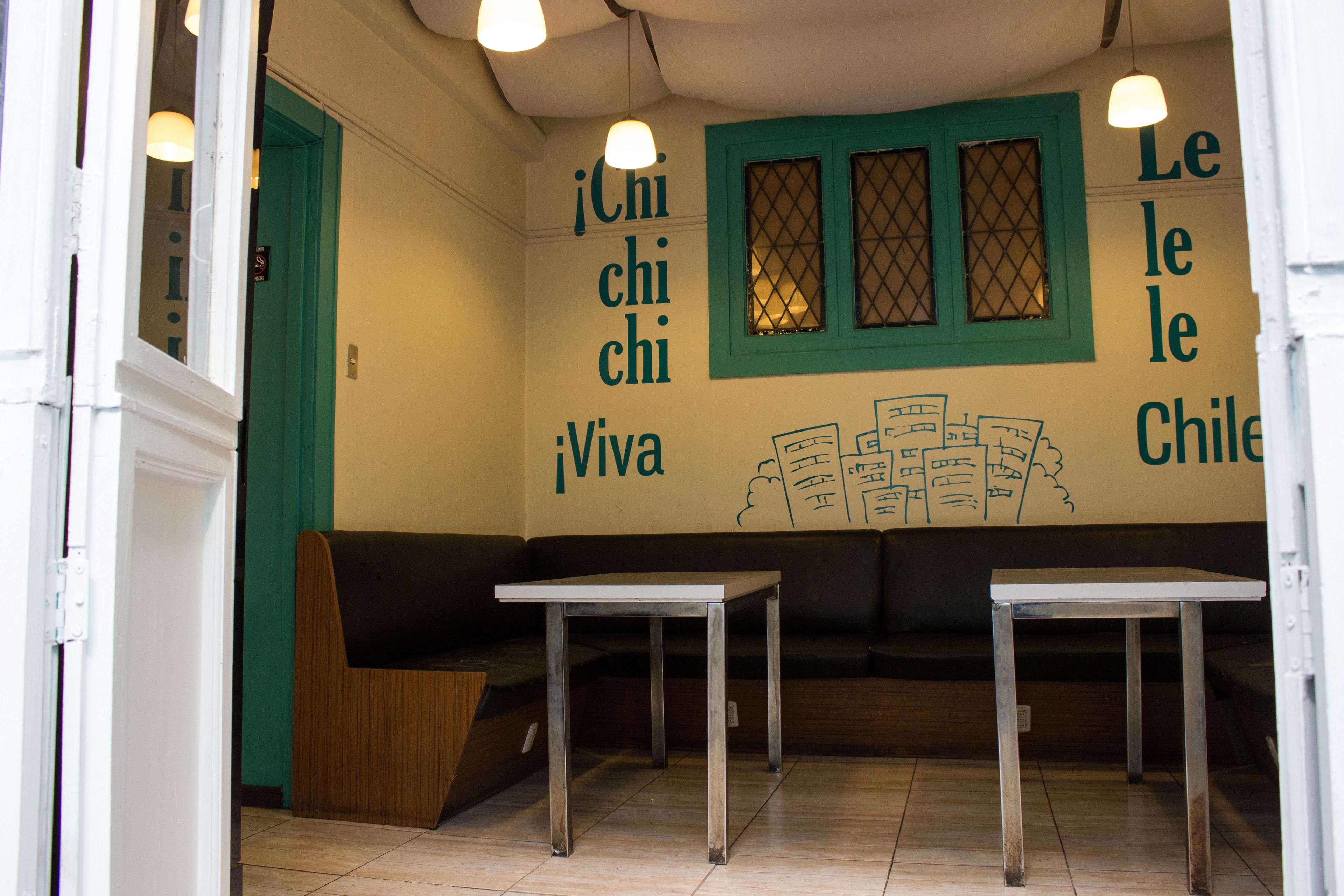 Interior de un restaurante con una puerta verde, dos mesas y una pared con texto e ilustraciones.
