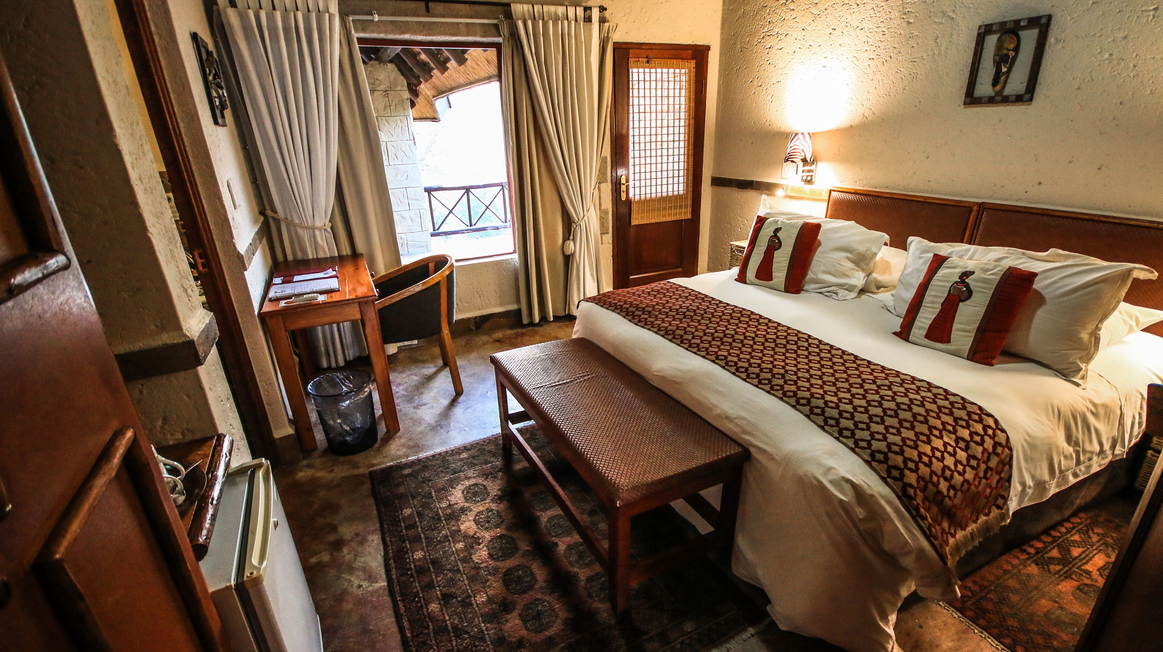 A bedroom with a bed, a desk, a chair, a trash bin, and a window with curtains.