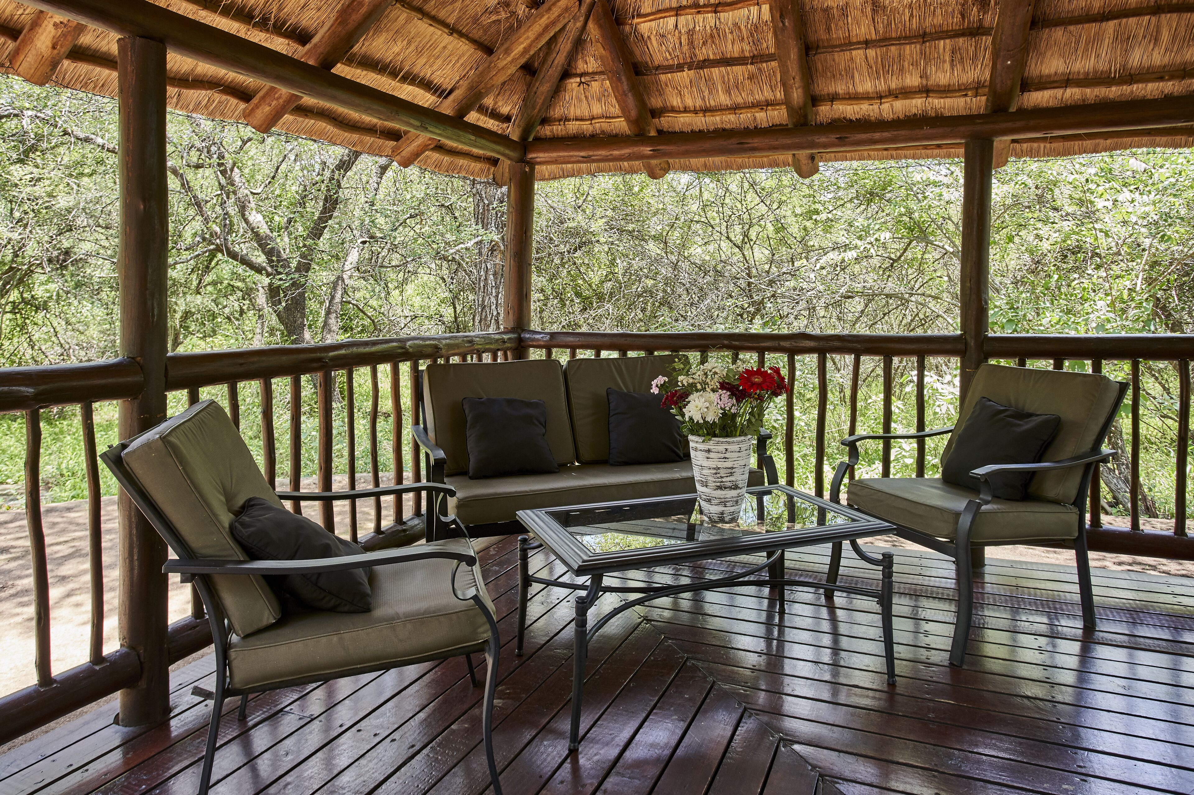 An outdoor wooden deck with a thatched roof, furnished with a sofa and two chairs, and a table with a vase of flowers.