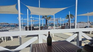 A beachside terrace with wooden tables and chairs, a bottle on one table, and a view of the beach with umbrellas and people.