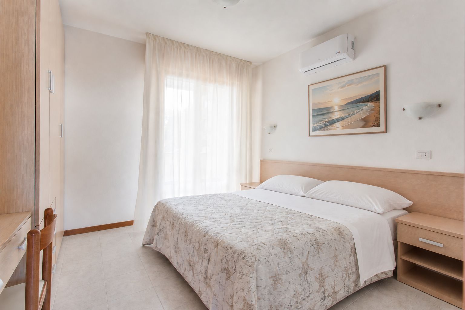 Habitación de hotel con cama, mesita de noche, escritorio, silla, ventana con cortinas, equipo de aire acondicionado y un cuadro en la pared.