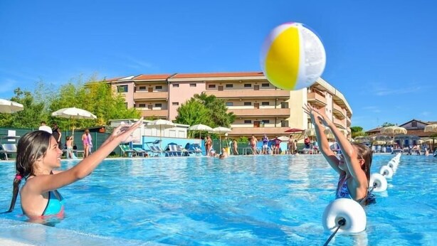 A swimming pool with people playing volleyball, a building with multiple balconies, and umbrellas.