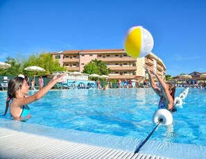 Una piscina con gente jugando al voleibol, un edificio con varios balcones y sombrillas.