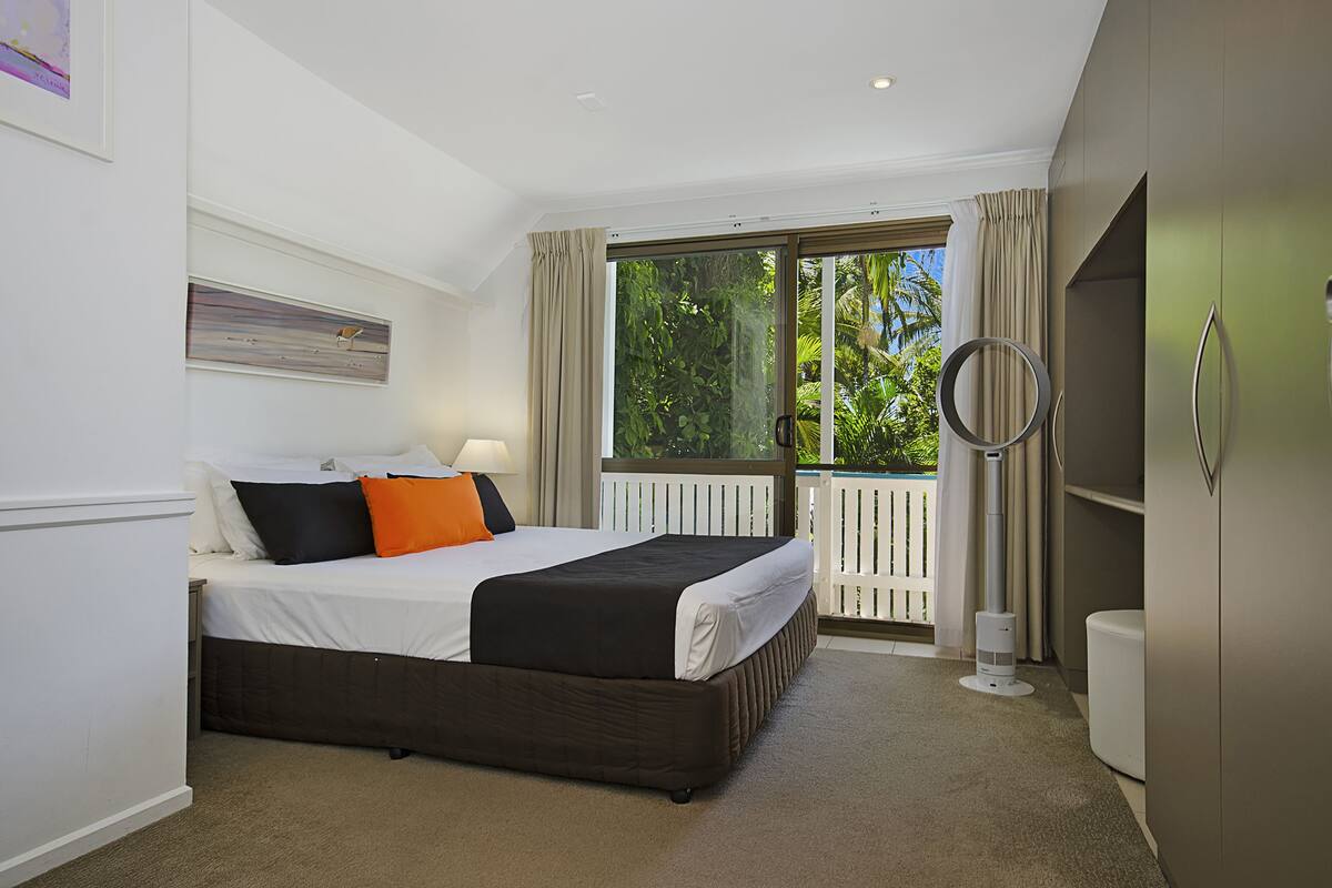 A bedroom with a bed, a nightstand, a lamp, a wardrobe, and a balcony with a view of greenery.