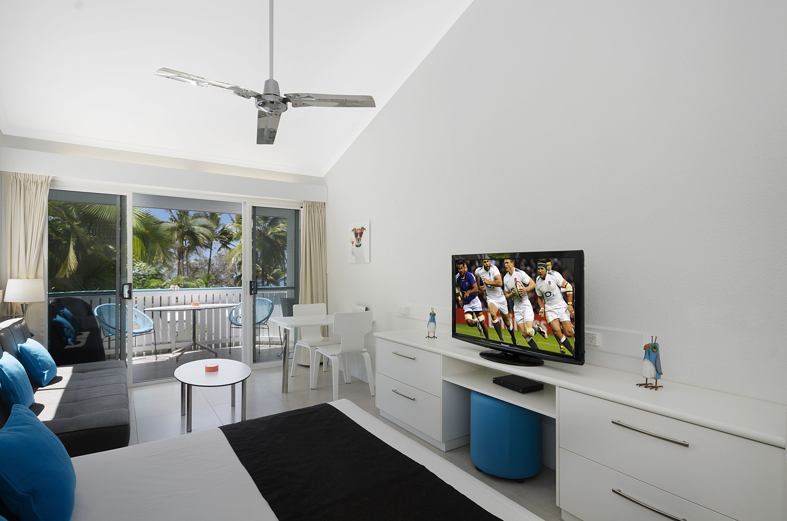 A modern hotel room with a large TV, white furniture, a balcony with a view of palm trees, and a ceiling fan.