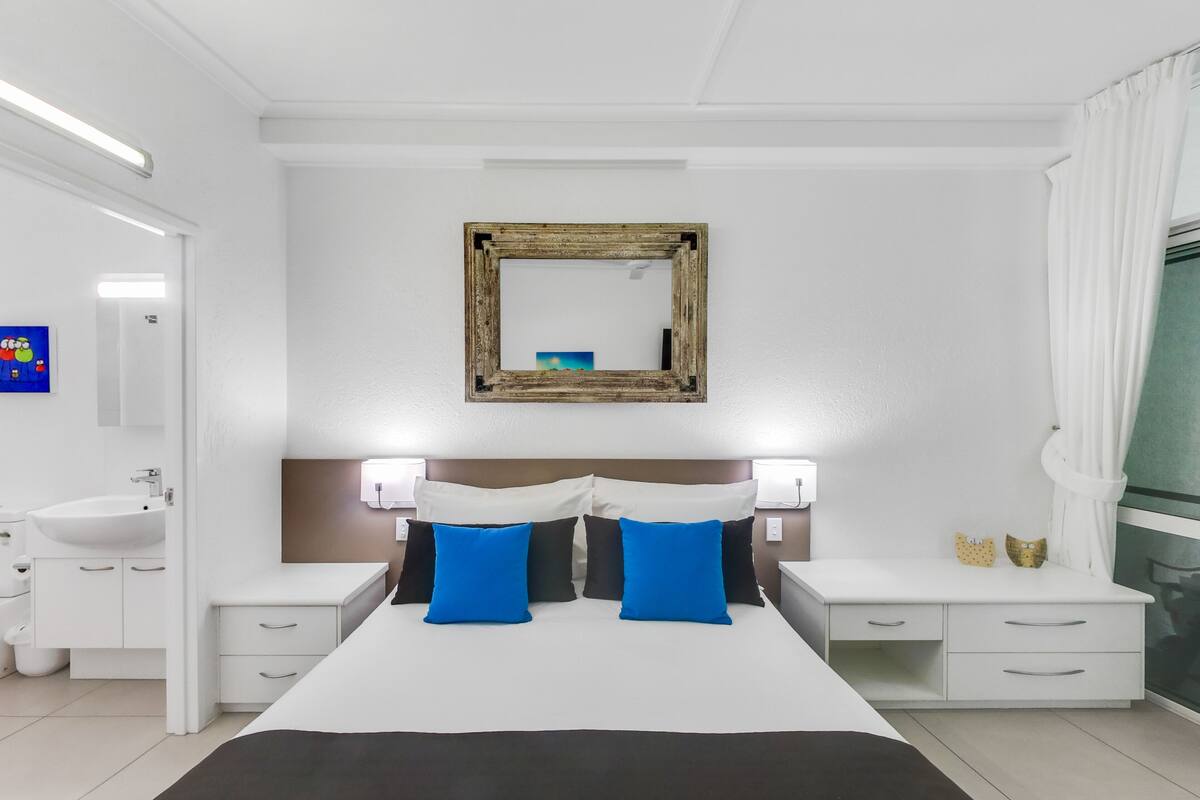 A modern bedroom with a large bed, bedside tables, a mirror, and a bathroom visible in the background.