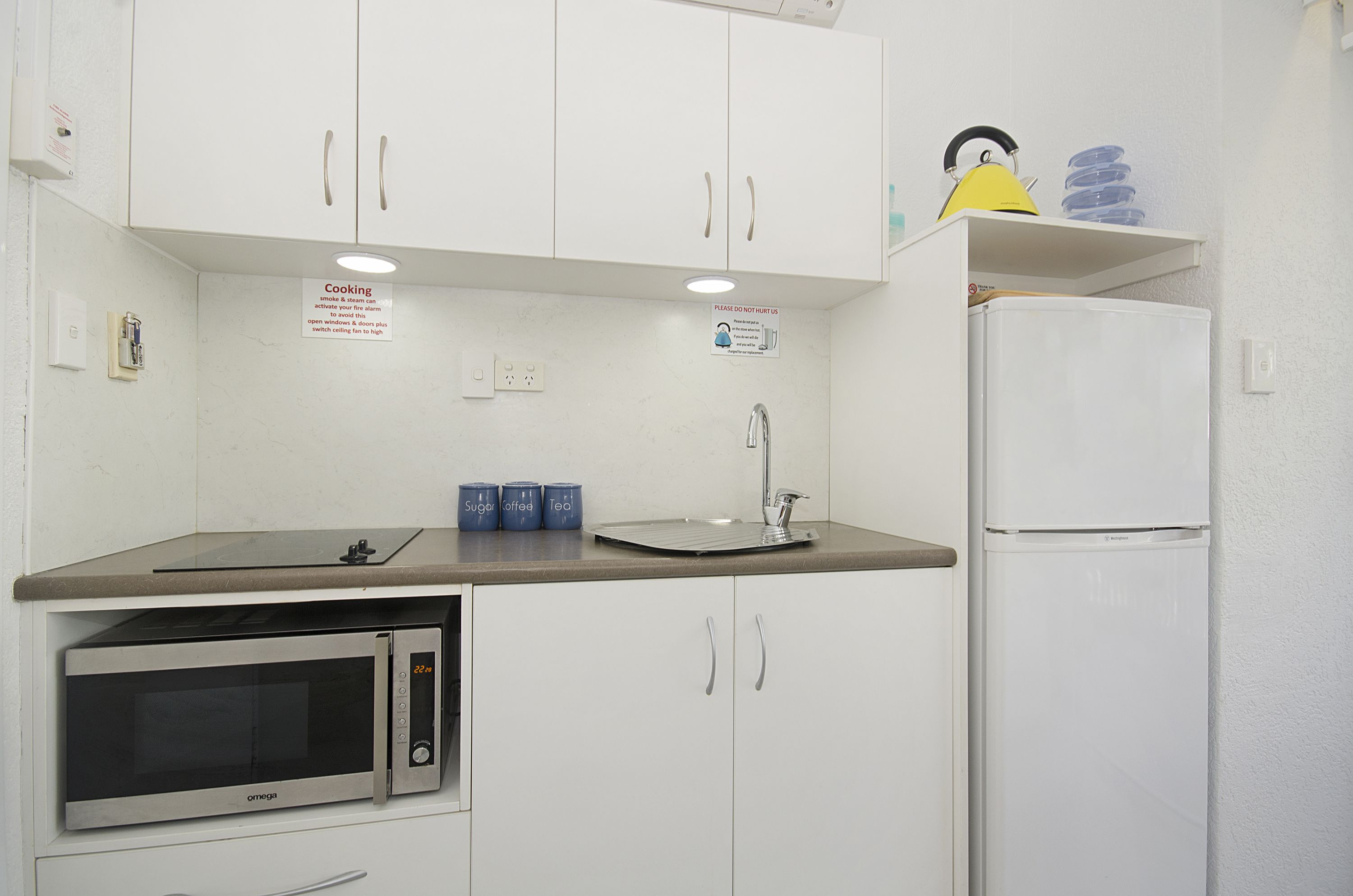 A compact kitchen with white cabinets, a microwave, a sink, and a refrigerator. There are three blue containers on the counter.