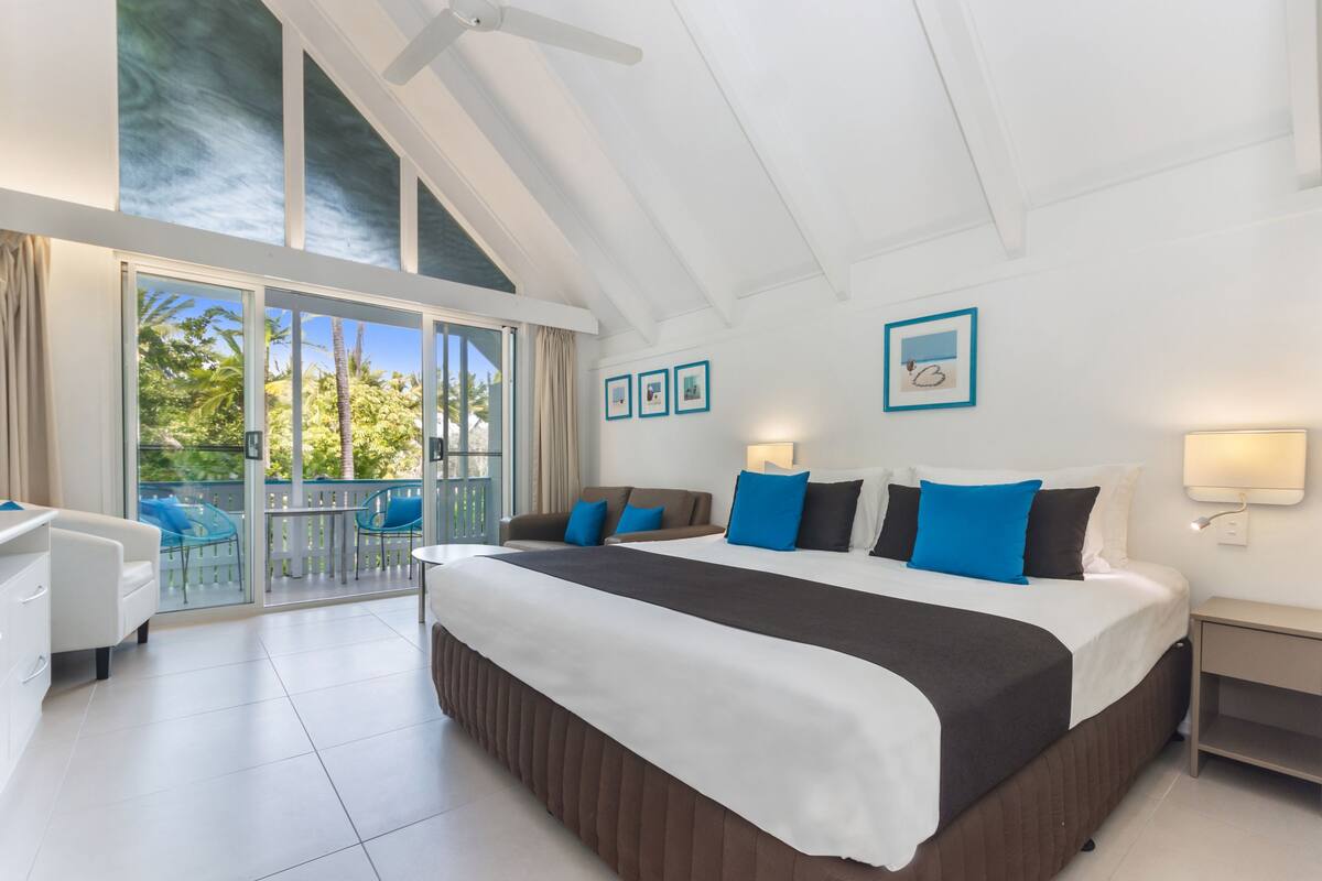 A spacious bedroom with a large bed, a balcony with chairs, and a ceiling fan.