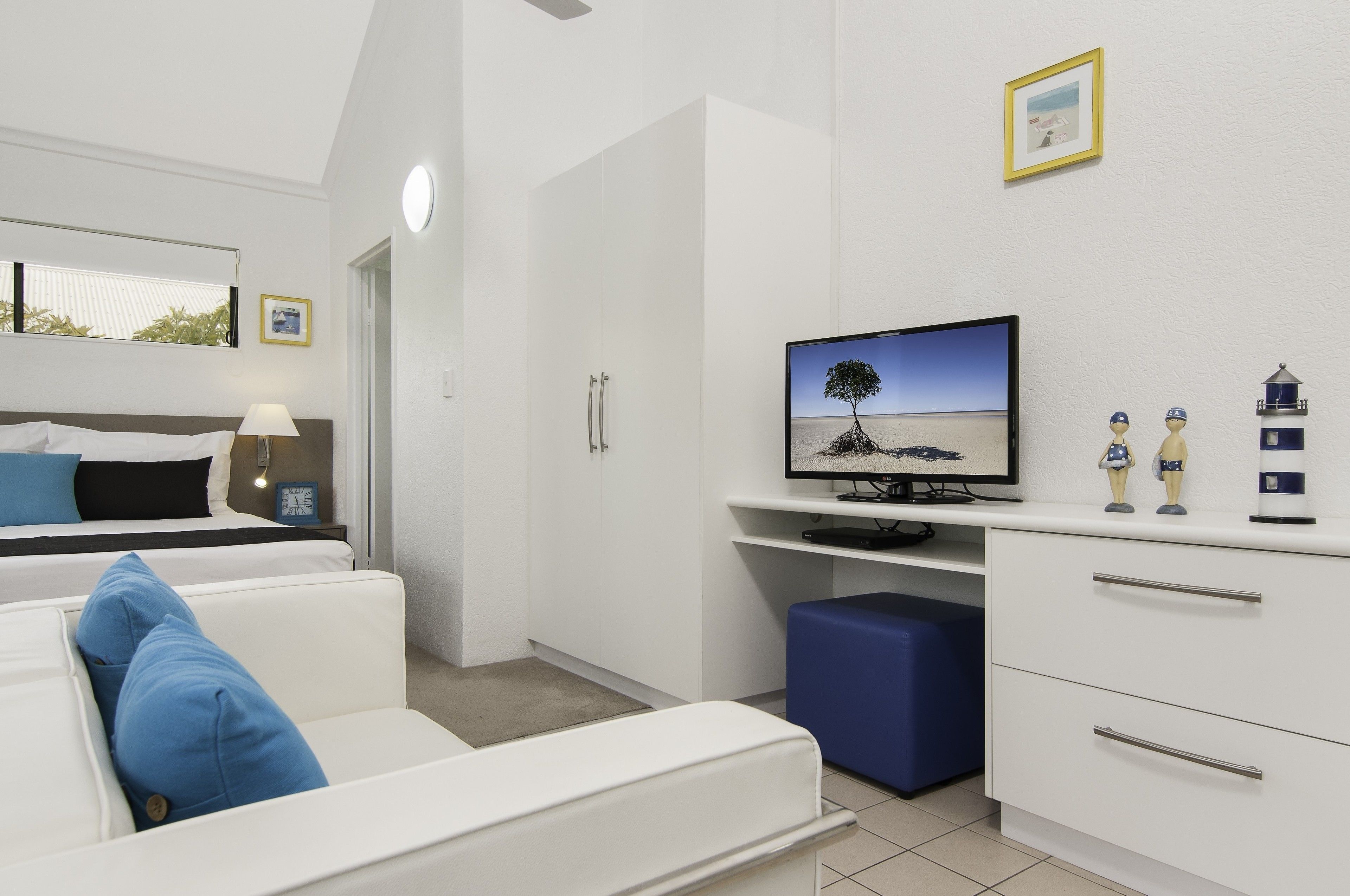 A modern hotel room with a bed, a white sofa with blue cushions, a television on a white stand, and a small desk with decorative items.