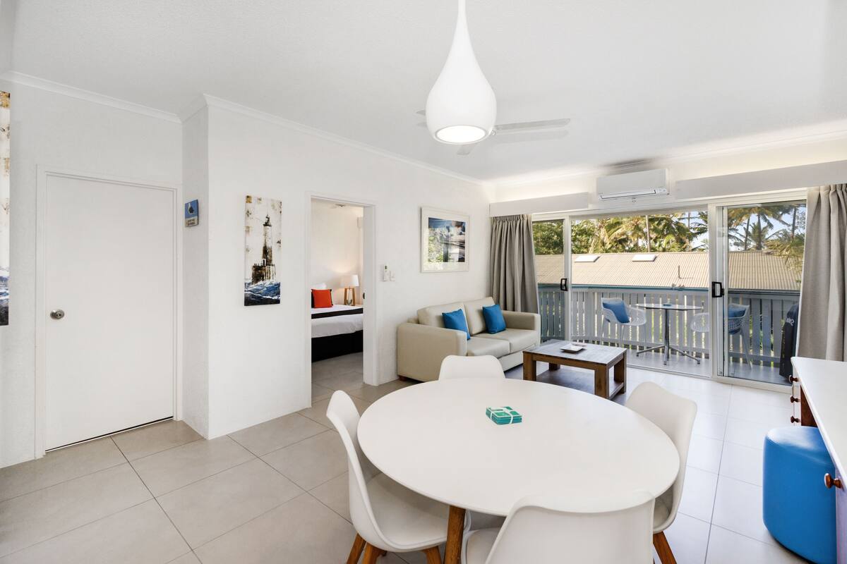 A modern living room with a round dining table, a sofa, and a balcony with a view of palm trees.