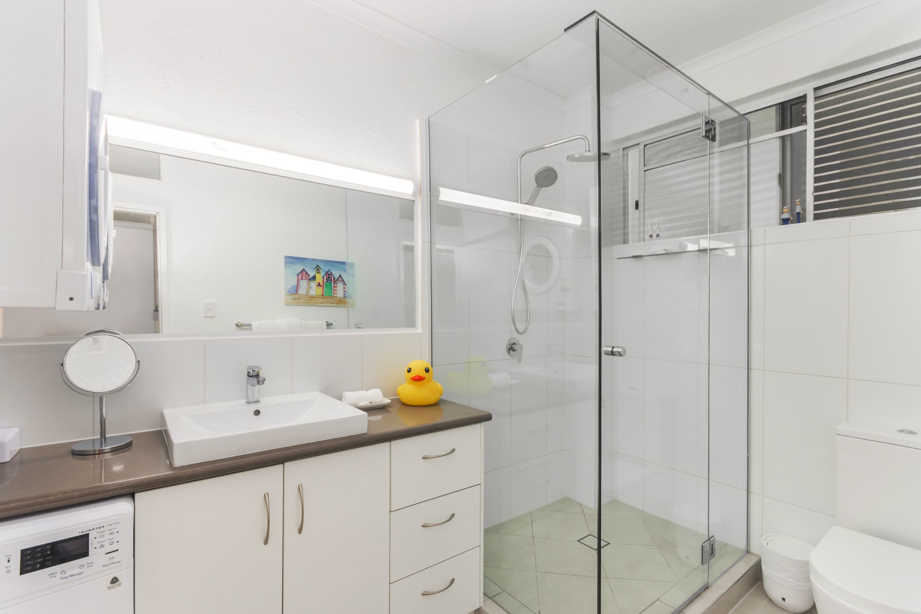 Un baño moderno con ducha de vidrio, un lavamanos blanco y un patito de goma pequeño en la encimera.
