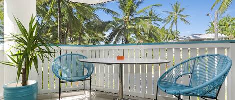 A balcony with a white table and two blue chairs, surrounded by white railings and palm trees.