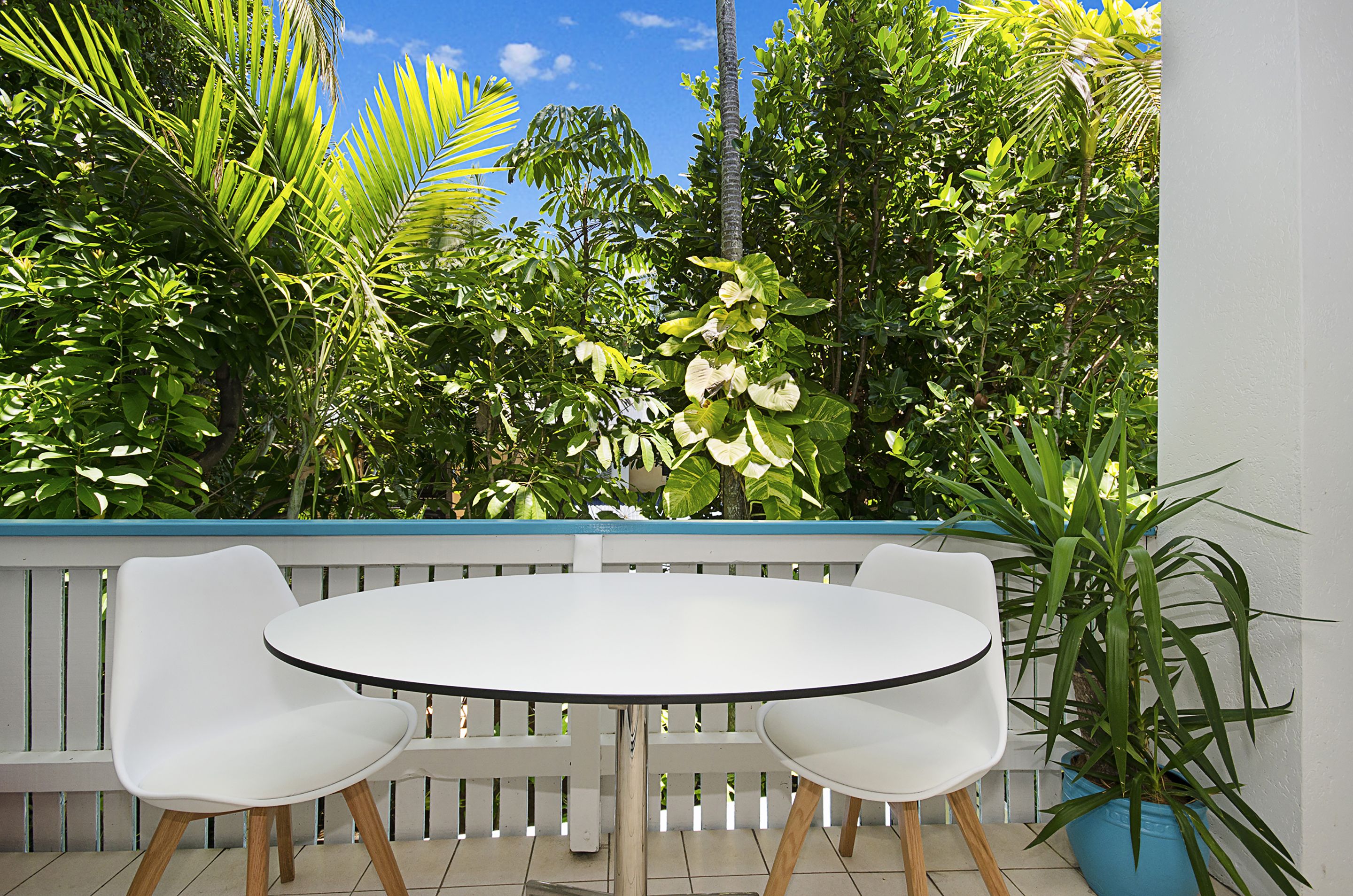 A round table with two white chairs, a white fence, and various green plants in the background.
