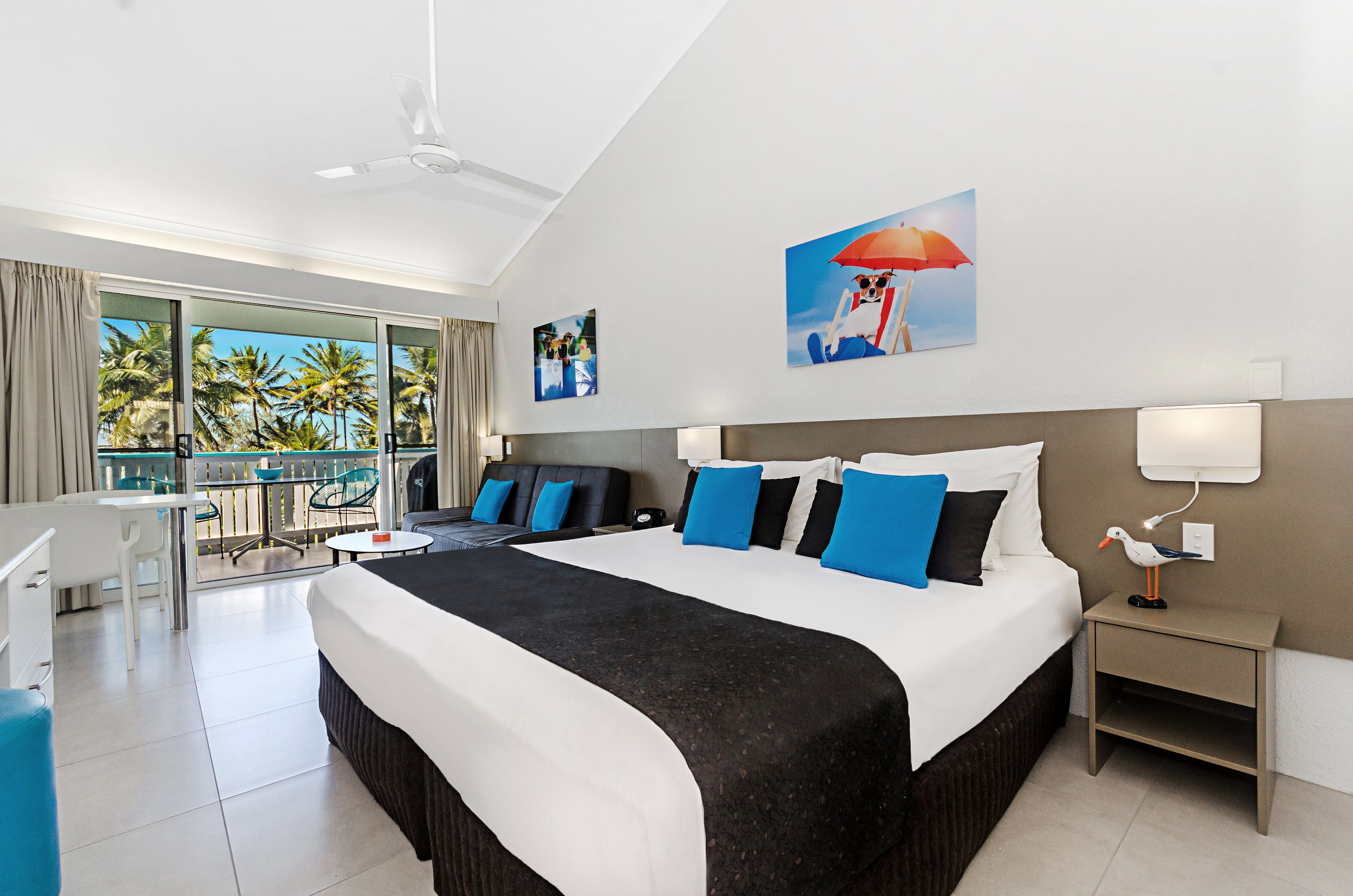 A hotel room with a large bed, a balcony with a view of palm trees, and a ceiling fan.