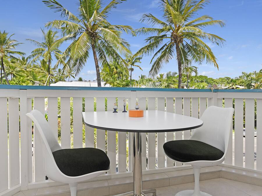 A white outdoor seating area with two chairs and a small table, surrounded by a white fence and palm trees in the background.