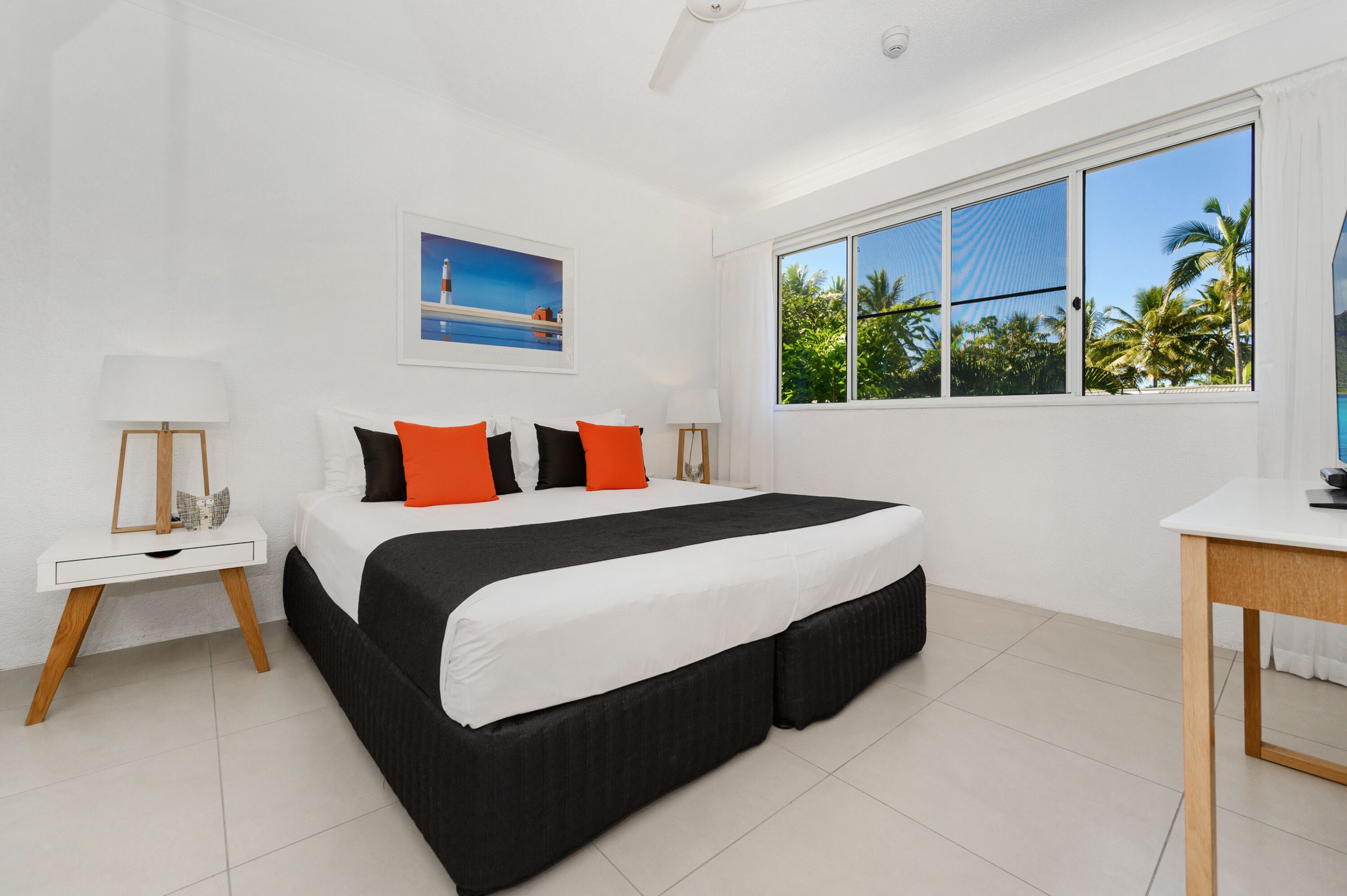 A bedroom with a large bed, white walls, a window with a view of palm trees, and a painting on the wall.