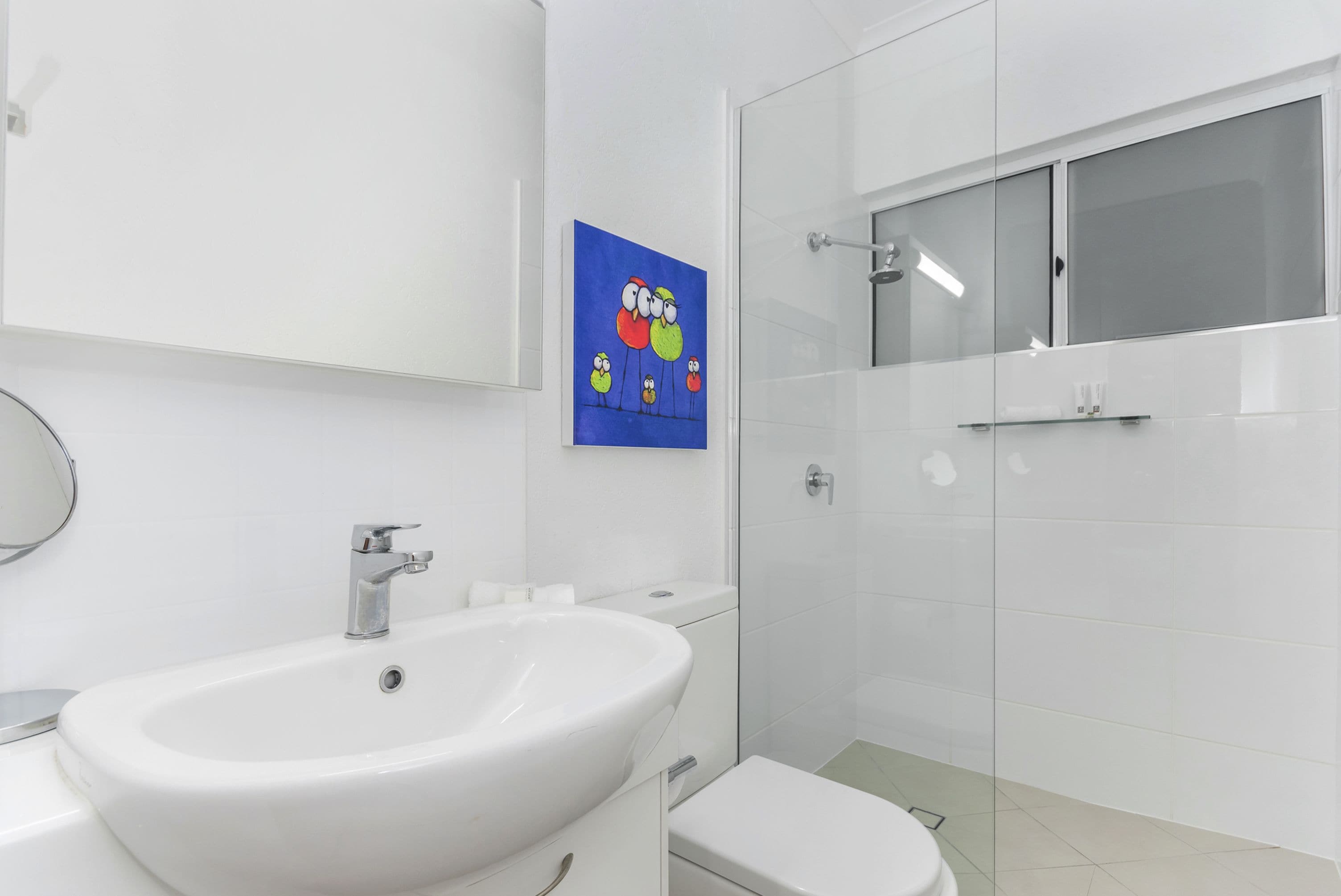 A modern bathroom with a white sink, toilet, and shower. There is a painting of birds on the wall.
