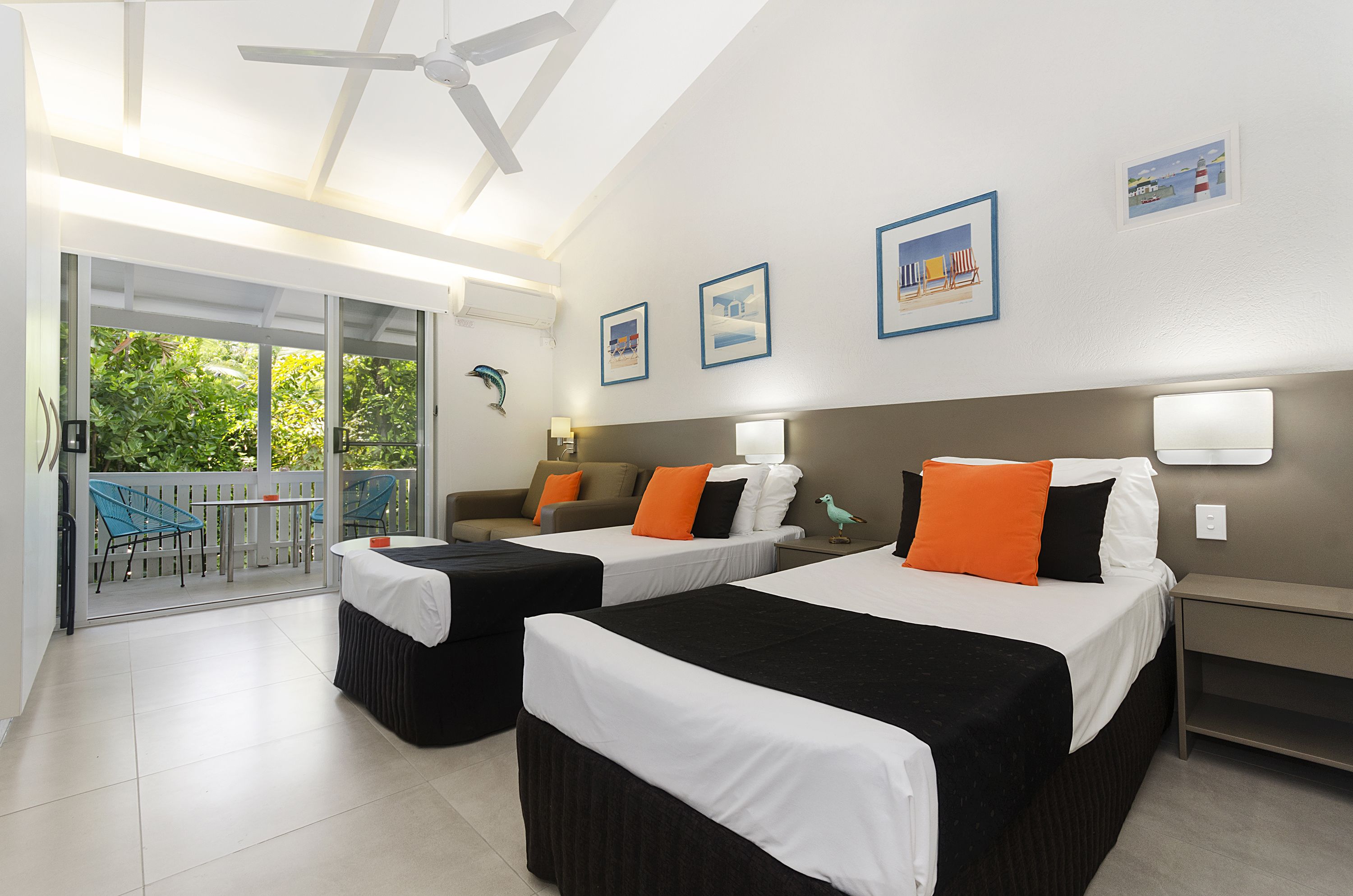A hotel room with two beds, a balcony with a view of greenery, and a ceiling fan.