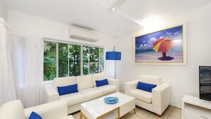 A modern living room with white sofas, blue cushions, a glass coffee table, and a large window with a view of greenery. - By The Sea Port Douglas (Port Douglas)