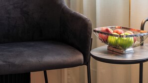 A room with a dark grey sofa, a small round table with a fruit bowl, a lamp, and a window with sheer curtains. - VICI Hotels Linköping (Linkoping)