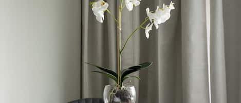 A hotel room with a bed, a chair, a small round table with a vase of white flowers, and a window with curtains.