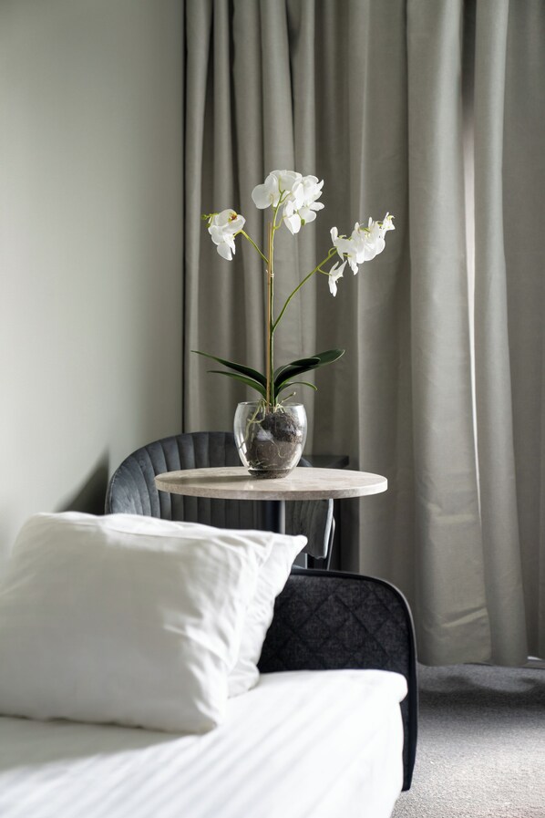 Una habitación de hotel con cama, una silla, una mesita redonda con un florero de flores blancas y una ventana con cortinas.