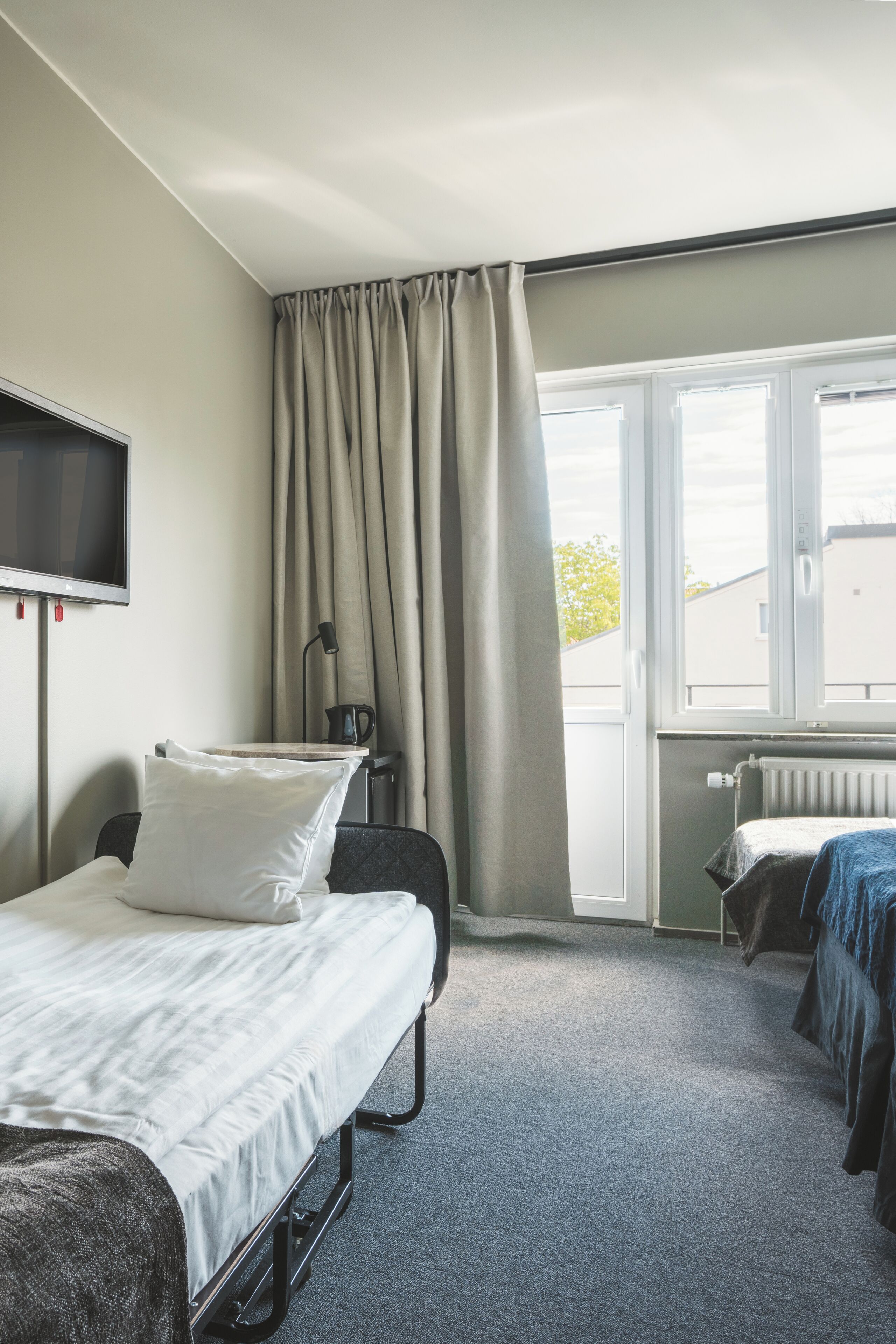 A hotel room with a bed, a desk, a chair, a TV, a window with curtains, and a radiator.