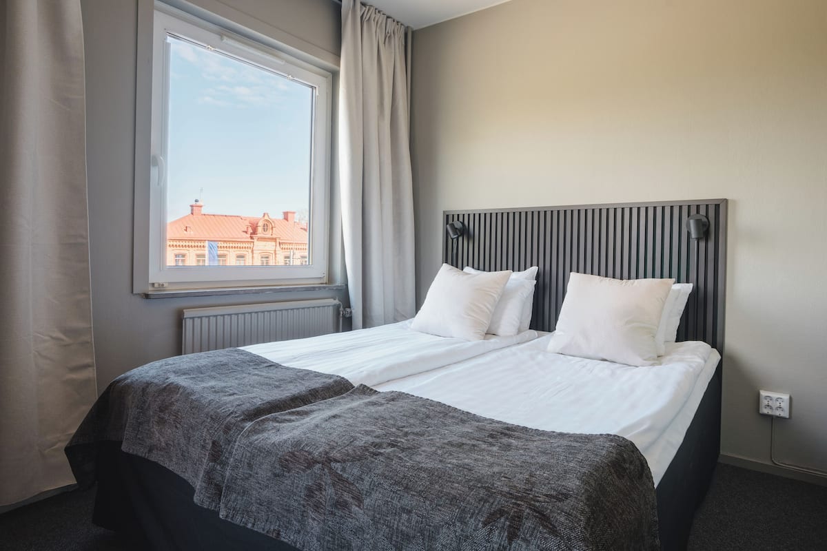 A hotel room with a large window, a bed with white linens, a grey headboard, and a grey throw blanket.