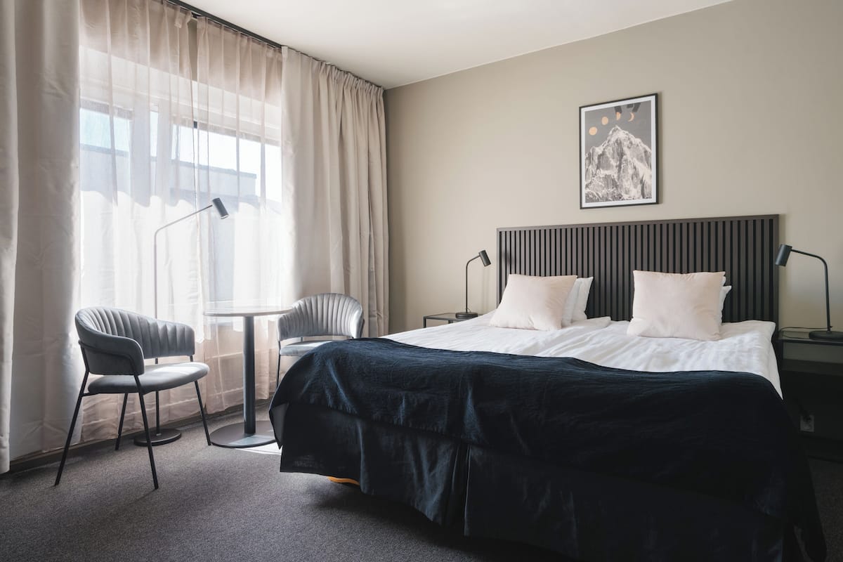 A hotel room with a bed, two chairs, a desk, and a large window with curtains.