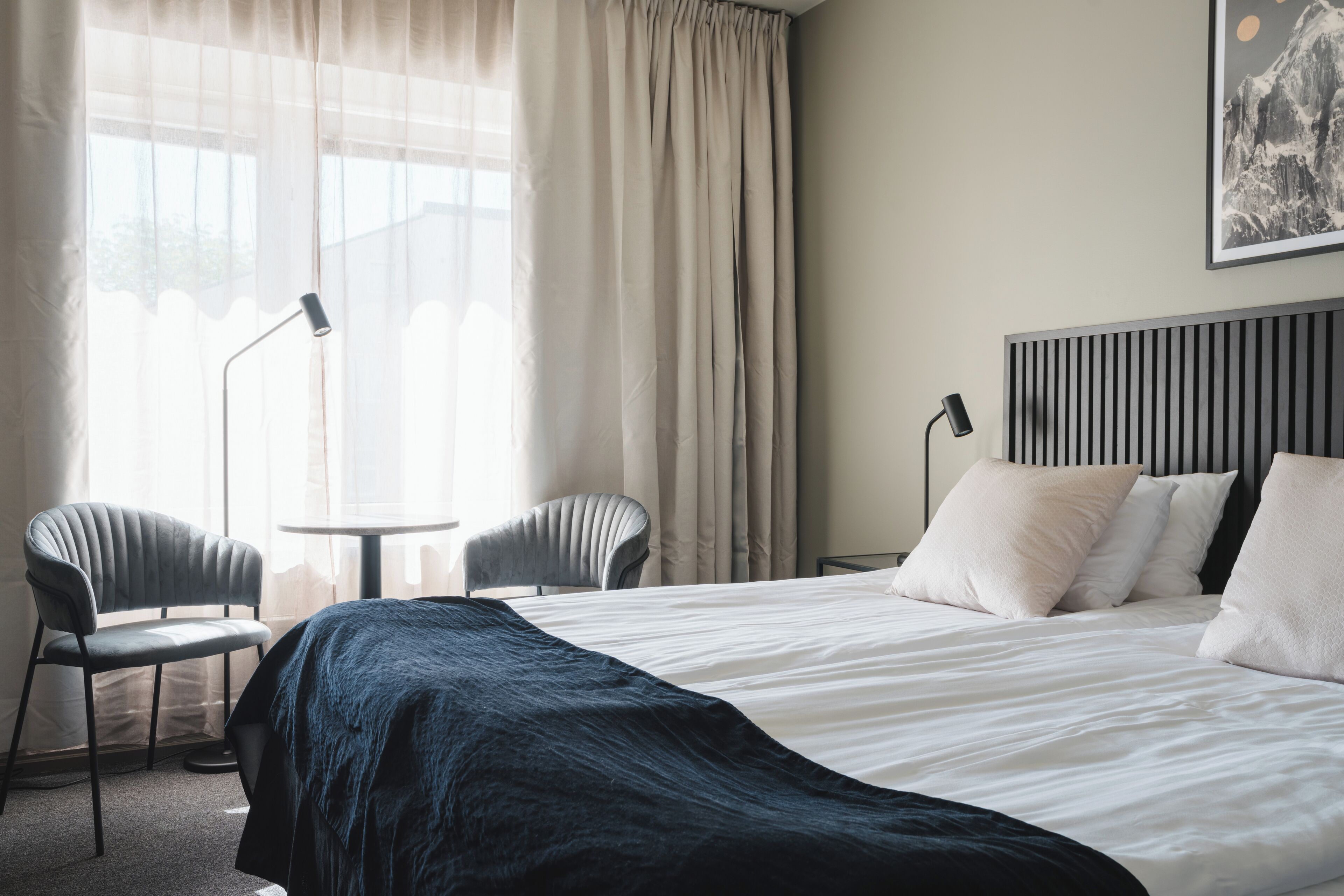 Una habitación de hotel con cama, dos sillas, un escritorio y un amplio ventanal con cortinas.