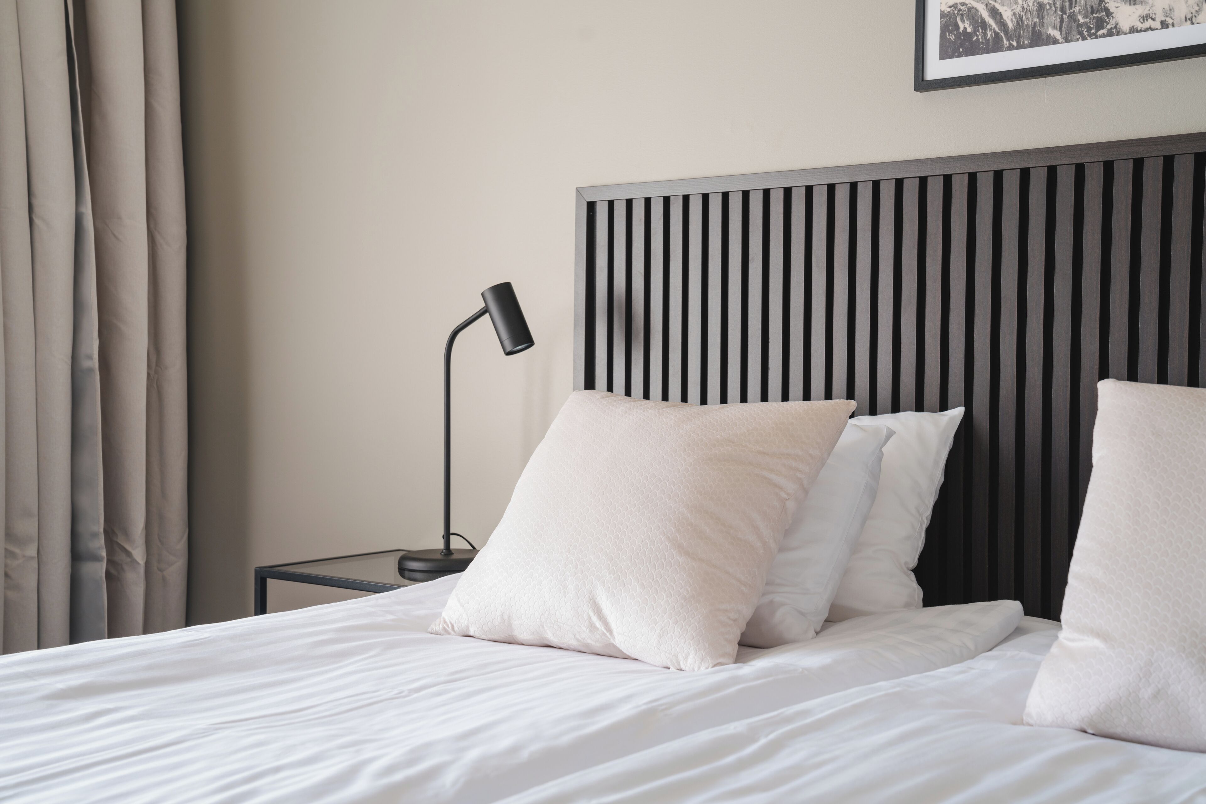 Una cama bien hecha con sábanas y almohadas blancas, una lámpara de noche negra y un cabecero moderno con líneas verticales.