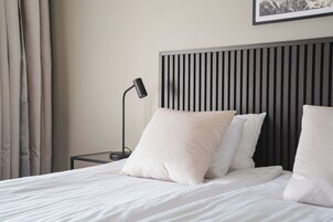 Una cama bien hecha con sábanas y almohadas blancas, una lámpara de noche negra y un cabecero moderno con líneas verticales.