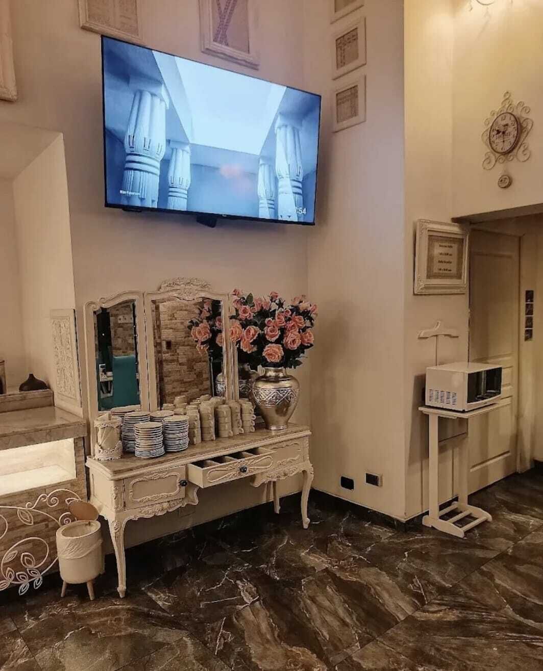 Een kamer met een aan de muur bevestigde televisie, een decoratieve console tafel met een bloemenvaas, en een witte kast met lades.