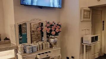 Een kamer met een aan de muur bevestigde televisie, een decoratieve console tafel met een bloemenvaas, en een witte kast met lades.