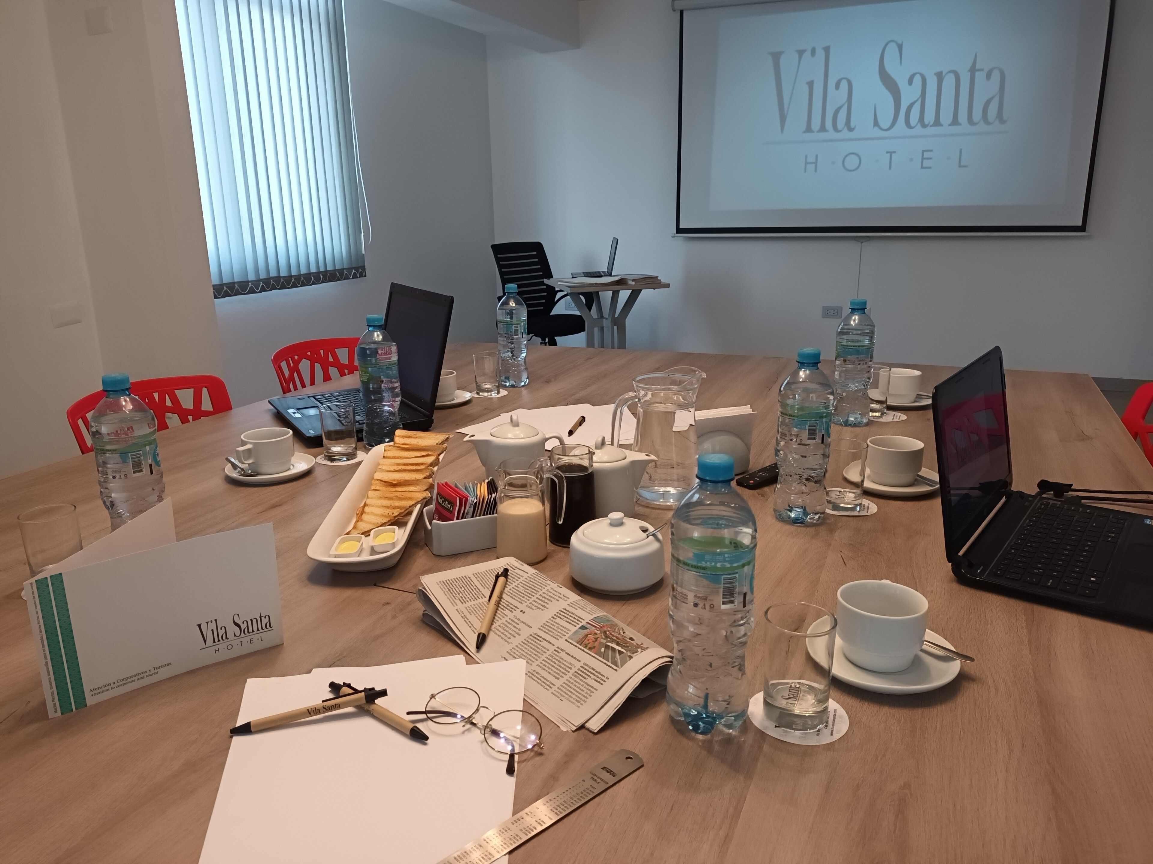 Uma sala de conferências no Vila Santa Hotel, com uma mesa de madeira preparada para uma reunião, laptops, garrafas de água e uma tela de projetor exibindo o nome do hotel.